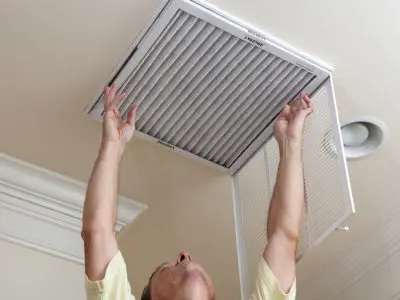 A person is reaching with both hands to replace their air filter on the ceiling.