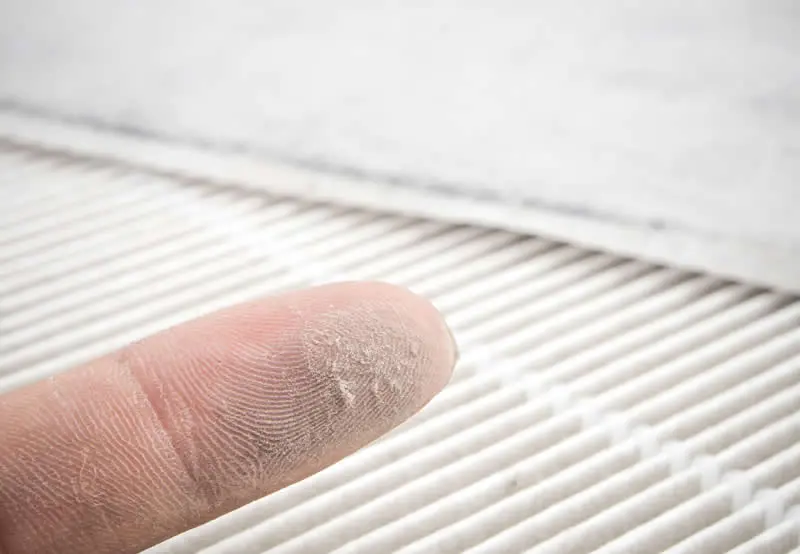 Image of a dirty air filter in the background. In front of it is a finger with dust on it, showing how the filter is dirty.