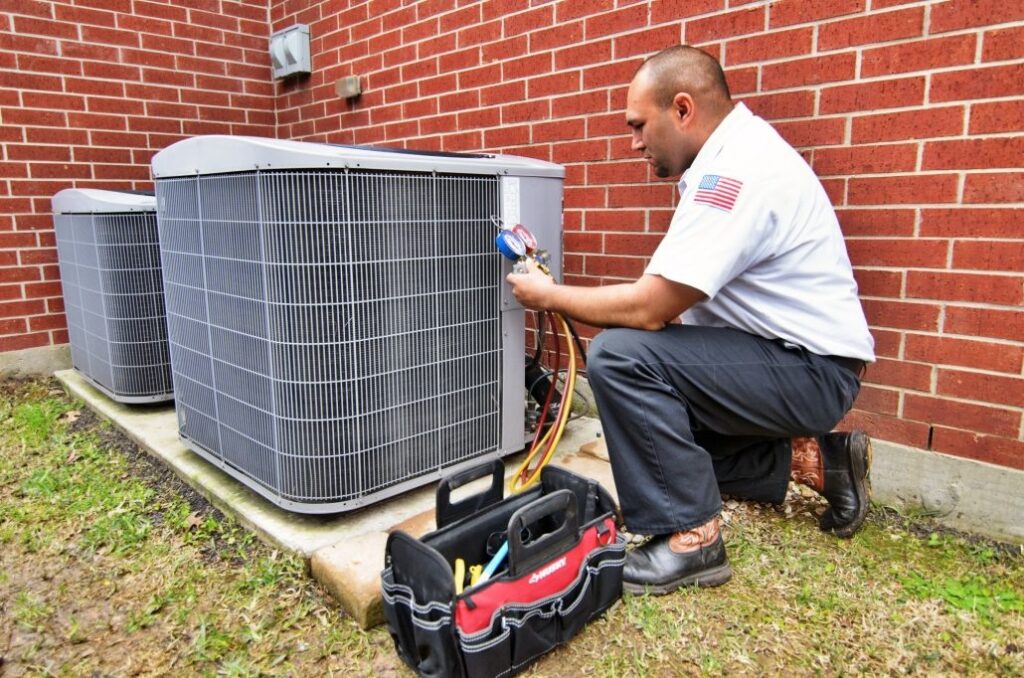 An AC technician is kneeling by an outdoor AC unit, performing a tune up.