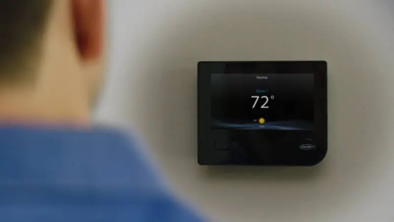 A smart thermostat is mounted on a wall with a man in the foreground, looking at it.