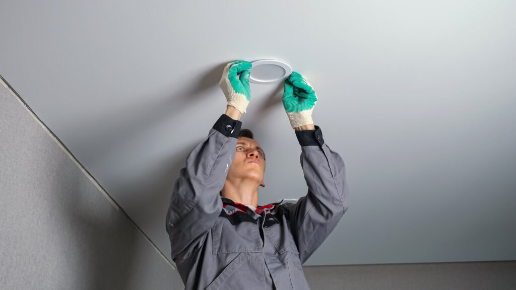 Electrician in work gloves and grey jacket installs light on ceiling standing on ladder during apartment renovation.