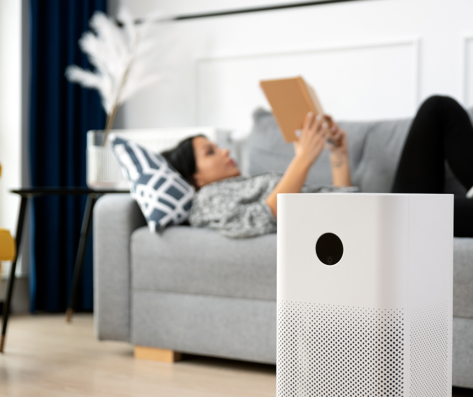 A woman in lying on her couch, reading a book, in the background. In front of her is an air scrubber.