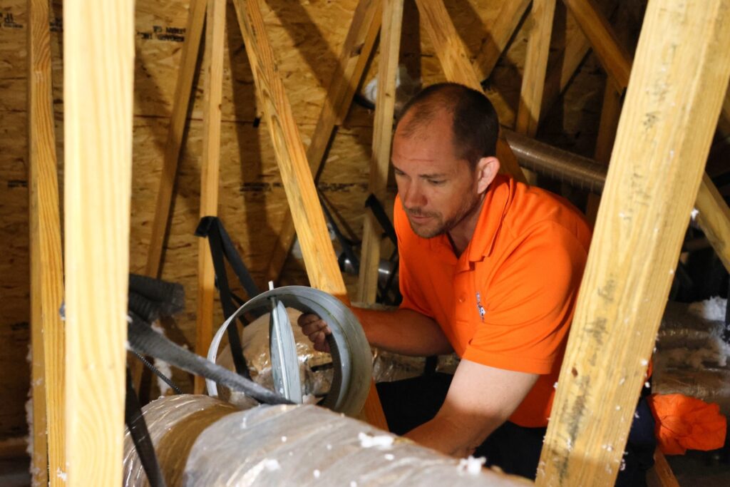 An HVAC technician is in an attic, repairing AC ductwork.